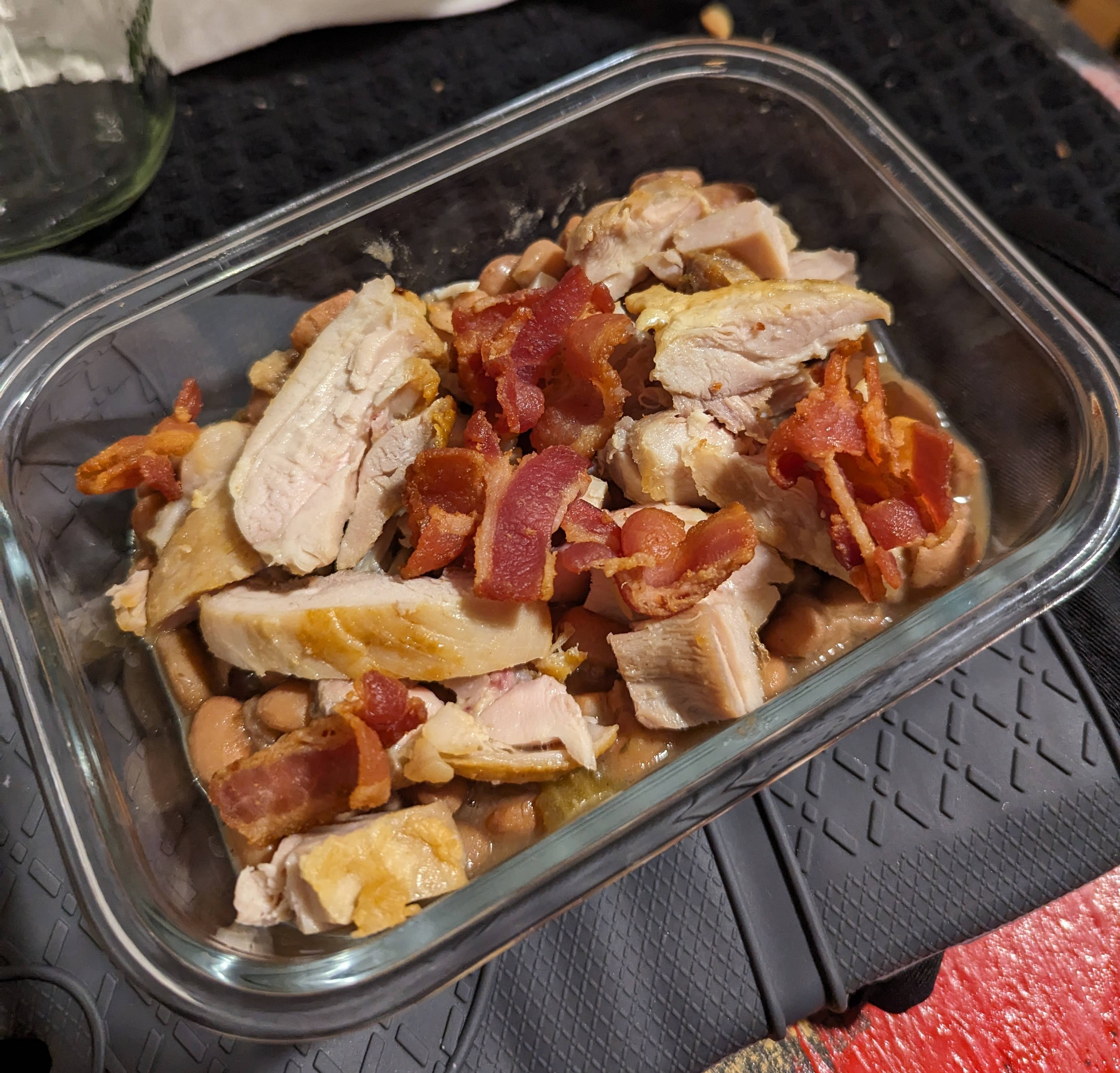 A glass container of beans with chicken and bacon on top.